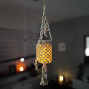 Könnte beinhalten: Eine hängende Makramee-Laterne mit warmem, leuchtendem Licht im Inneren. Die cremefarbene Laterne hat ein Rautenmuster und eine lange Quaste. Die Laterne ist an einem geknoteten Seil mit Holzperlen aufgehängt.
