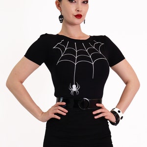 Puede incluir: Una camiseta negra con un diseño de araña y telaraña blanca. La camiseta tiene mangas cortas y un cuello redondo.