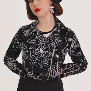 Peut inclure: Une veste de moto noire avec un motif d'araignées et de toiles d'araignées blanches. La veste présente une fermeture éclair asymétrique argentée sur le devant, un large col et des poignets zippés, portée sur un haut blanc à col noir.