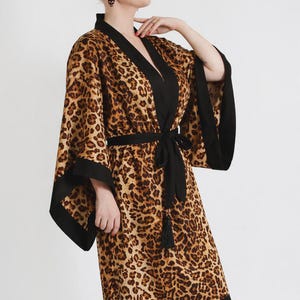 Op de afbeelding: Een luipaardprint badjas met zwarte afwerking en bijpassende riem. De badjas heeft een klassieke kimono-stijl met wijde mouwen en een zoom tot op de knie. De bruine en zwarte dierenprint wordt gecontrasteerd door de effen zwarte accenten.