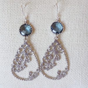 May include: A pair of silver-coloured drop earrings. Each earring has a faceted, round, blue gemstone at the top, connected to a teardrop-shaped filigree design. The filigree has an ornate, openwork pattern.