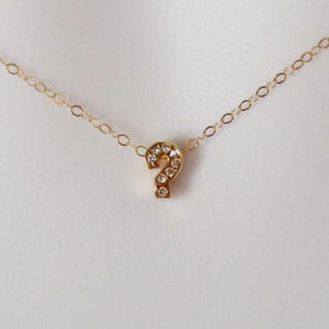 Könnte beinhalten: Eine zarte, goldfarbene Halskette mit einem kleinen Fragezeichen-Anhänger. Der Anhänger ist mit kleinen, klaren Edelsteinen besetzt, die für Glanz sorgen. Die Kette hat ein feines Kettenglied-Design und wirkt minimalistisch und elegant.