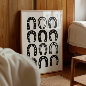 Puede incluir: Un cuadro enmarcado con ilustraciones de herraduras negras sobre fondo blanco. Las herraduras presentan diseños decorativos variados, como estrellas y puntos. El marco es de color madera clara.