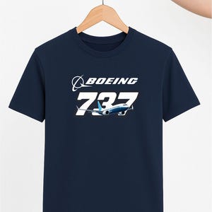 May include: Navy blue t-shirt with the Boeing logo and '737' in white, plus an image of a 737 aircraft. The t-shirt is displayed on a wooden hanger. The design is a tribute to aviation.