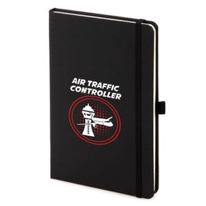 Puede incluir: Cuaderno negro con el texto "AIR TRAFFIC CONTROLLER" encima de un círculo rojo con un gráfico blanco de un avión y una torre de control. El cuaderno tiene un cierre de banda elástica negra.