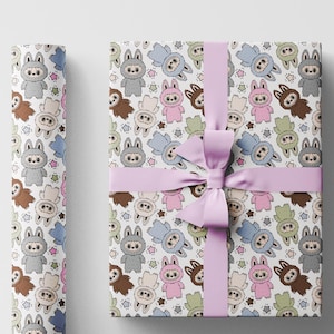 Puede incluir: Caja de regalo envuelta y rollo de papel de regalo con un patrón repetido de conejitos de dibujos animados en varios colores. La caja está atada con una cinta morada clara. El fondo es blanco.