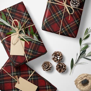 May include: Overhead shot of wrapped gifts with red plaid paper, tied with twine and adorned with sprigs of greenery and pinecones. A pair of scissors and a spool of twine are also present, suggesting a holiday or special occasion theme.