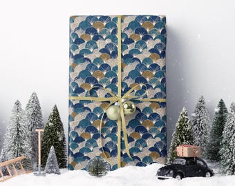 May include: A wrapped gift with a blue and gold fan pattern, tied with a gold ribbon and a gold ornament. Miniature snow-covered pine trees and a black toy car carrying a gift box are in the foreground, creating a festive holiday scene.