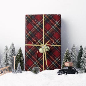 May include: A large gift wrapped in red plaid paper with a gold ribbon and ornaments. Miniature snow-covered pine trees and a black toy car carrying a gift box are in the foreground, creating a festive holiday scene.