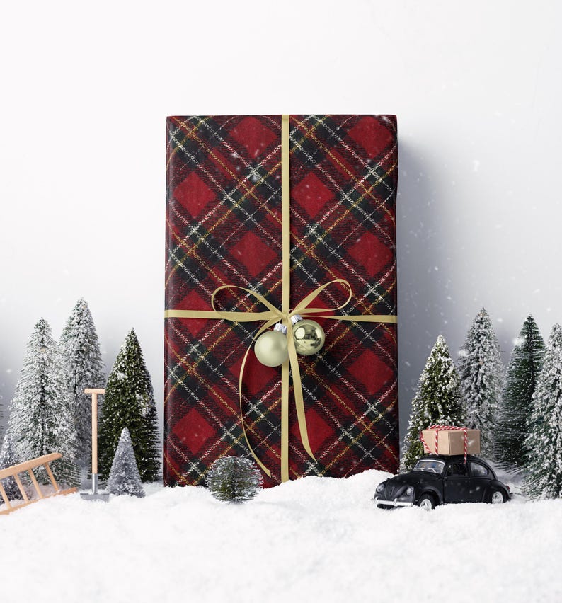 May include: A large gift wrapped in red plaid paper with a gold ribbon and ornaments. Miniature snow-covered pine trees and a black toy car carrying a gift box are in the foreground, creating a festive holiday scene.