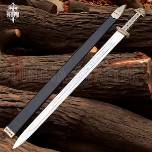 May include: A silver sword with a decorative handle and a black sheath. The handle is adorned with red and gold accents. The blade has etched text. The sword is displayed on a wooden surface.
