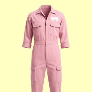 Pode incluir: Macacão rosa claro com gola, dois bolsos no peito e dois bolsos inferiores. O macacão tem mangas três quartos e cinto na cintura. Dois patches brancos em forma de coração estão no peito esquerdo.