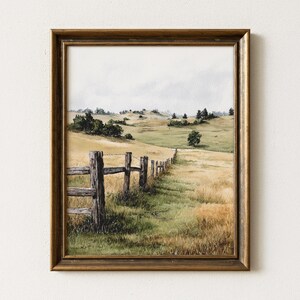 May include: Watercolor landscape art print in a gold-toned wooden frame. The artwork depicts a rural scene with a wooden fence, rolling hills, and a field of tall grass. The color palette includes shades of green, yellow, and brown.