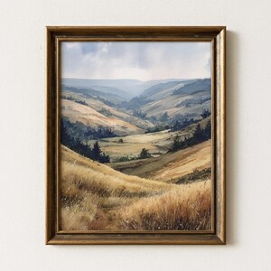 Puede incluir: Pintura de paisaje enmarcada que representa colinas y valles en tonos dorados, verdes y azules. La obra de arte presenta un estilo de acuarela detallado con una paleta de colores cálidos y naturales. El marco es de color oro antiguo.