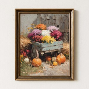May include: A framed painting shows a blue wooden cart filled with colourful chrysanthemums. Several orange pumpkins and gourds are scattered around the cart, with a weathered wooden fence in the background. The artwork has a warm, autumnal feel.
