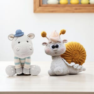 May include: Two handmade crochet stuffed animals: a gray hippo wearing a blue hat and striped shirt, and a snail with a yellow shell and pink cheeks. The hippo is seated, and the snail is curled up. Both have black eyes.