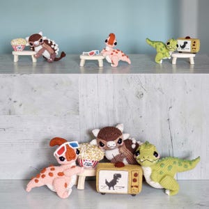 May include: Crocheted dinosaur figurines in various colors, including brown, pink, and green. Some dinosaurs are posed with popcorn, 3D glasses, and a miniature television set. The TV displays a dinosaur silhouette.