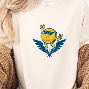 May include: Cream-colored t-shirt featuring a cartoon coin character wearing sunglasses and making a peace sign. The coin is yellow with a blue and yellow wing design. The shirt is a casual style.