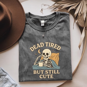May include: Dark gray t-shirt with the text "DEAD TIRED BUT STILL CUTE" and a cartoon skeleton design. The skeleton is sitting up in bed, holding a mug. A brown hat and necklace are also in the image.