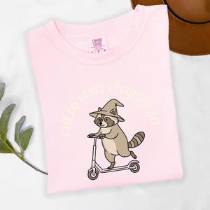 May include: A light pink t-shirt featuring a graphic of a raccoon wearing a pointed hat and riding a scooter. The text "Off to start chapter 30" is printed in a light cream color in an arch above the raccoon illustration on the front of the shirt.
