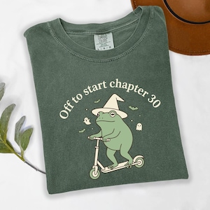 May include: A forest green t-shirt featuring a graphic of a light green frog wearing a wizard hat and riding a scooter. The text above the frog reads "Off to start chapter 30" in a cream color, accompanied by small illustrations of ghosts and bats.