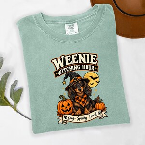 May include: A sage green t-shirt featuring a graphic of a long-haired dachshund wearing a witch hat and an orange patterned bandana. The design includes two pumpkins, a full moon with bats, and the text "WEENIE WITCHING HOUR" and "Long Spooky Loved".