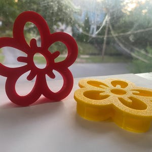 May include: Two flower-shaped cookie cutters. One is red and stands upright, while the other is yellow and lies flat. The cutters are made of plastic and have a simple, open design, perfect for baking.