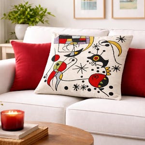 Può includere: Un divano bianco con due cuscini rossi e un cuscino decorativo con un design astratto in nero, rosso, giallo e blu. Una candela accesa e una piccola pianta sono su un tavolo vicino.