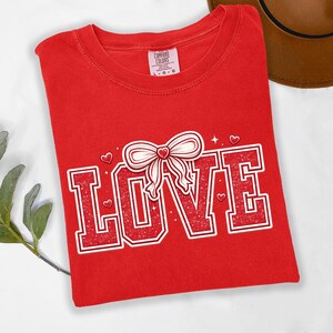 May include: A red t-shirt with the word "LOVE" in a distressed, outlined font. A white bow with a red heart is above the "O". Small hearts and stars are scattered around the word. The shirt has a tag that says "Comfort Colors".