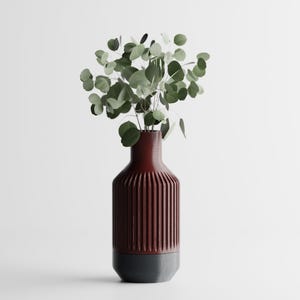 Könnte beinhalten: Eine weinrote und graue Vase mit geripptem Design, die ein Bündel grüner Eukalyptusblätter enthält. Die Vase hat eine zylindrische Form mit einem dunkelgrauen Sockel. Der Eukalyptus hat runde, grüne Blätter.