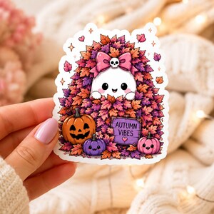May include: A die-cut sticker featuring a white ghost with a pink bow and skull accent, nestled in a pile of purple and orange autumn leaves. The design includes an orange jack-o'-lantern, a purple pumpkin, a pink pumpkin, and a sign reading AUTUMN VIBES.