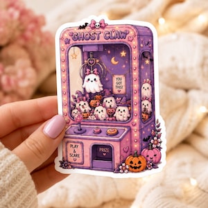May include: A die-cut sticker of a purple and pink claw machine labeled GHOST CLAW. The machine contains white ghost figures, small pumpkins, and a sign reading YOU GOT THIS. The control panel features a joystick and buttons labeled PLAY & SCARE.