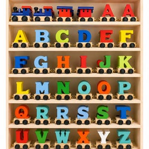 May include: A wooden display shelf containing a collection of toy train carriages, each carrying a single letter of the alphabet. The letters are painted in various bright colours including red, blue, yellow, green, orange, and light blue, with black wheels.