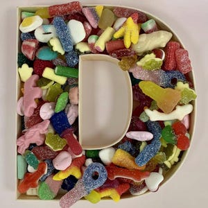 Puede incluir: Una caja de madera con forma de la letra "D" llena de una variedad de caramelos de goma coloridos. Los caramelos incluyen formas rojas, amarillas, azules y verdes, algunos con cobertura de azúcar. El fondo es rosa claro.