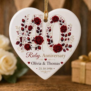 Puede incluir: Un adorno de cerámica blanco en forma de corazón con el número 40 decorado con rosas rojas y hojas verdes. El texto dice Ruby Anniversary, Olivia & Thomas y 25.10.1986. Cuelga de un cordón dorado sobre un fondo de madera desenfocado.