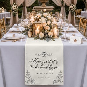 May include: A cream-colored fabric table runner featuring the black printed text How sweet it is to be loved by you, the names Emily & James, and the date October 24, 2026. The design includes a line drawing of two interlocking rings and floral vines.