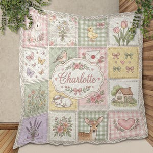 Könnte beinhalten: Eine Steppdecke mit einem Raster aus pastellfarbenen Quadraten, die mit Illustrationen von einem Hasen, einer Ente, einem Reh, einem Haus, Blumen, Schmetterlingen und Herzen versehen sind. Ein zentrales ovales Feld zeigt den Namen Charlotte.