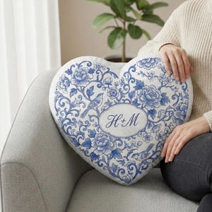 Peut inclure: Coussin décoratif en forme de cœur avec un motif floral bleu et blanc. Le coussin présente un oiseau et les initiales "H+M" dans un ovale. Le coussin est sur une chaise grise.