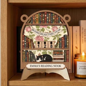 May include: Wooden bookshelf-shaped reading tracker with the text "BOOKS READ THIS YEAR" and "EMMA'S READING NOOK". It features a black and white cat, a floral background, and a counter showing "128".