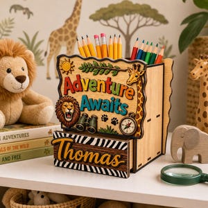 May include: A wooden desk organiser with the words "Adventure Awaits" and the name "Thomas". The organiser features a lion, giraffe, compass, and binoculars design. Pencils are inside the organiser. A stuffed lion, giraffe figurine, and books are also present.
