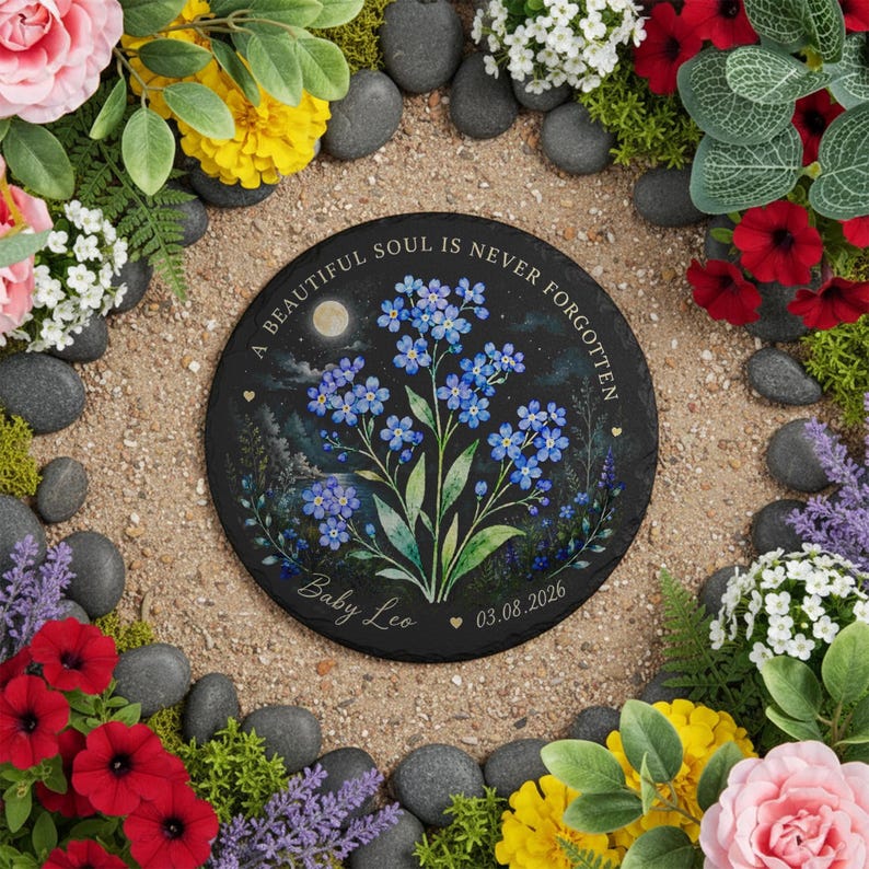 May include: A round black memorial stone with the text "A BEAUTIFUL SOUL IS NEVER FORGOTTEN" and "Baby Leo 03.08.2026". The stone features a watercolor painting of blue forget-me-nots under a moonlit sky. The stone is surrounded by flowers and pebbles.