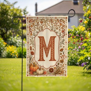 May include: A rectangular garden flag featuring a large, rust-colored letter M centered within a decorative border. The design includes autumn-themed illustrations of orange pumpkins, green gourds, brown corn, and various fall flowers and foliage.