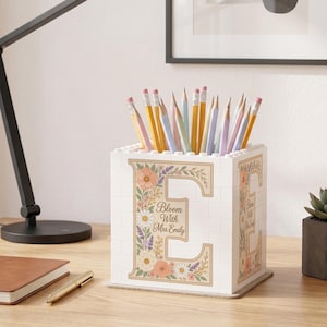 Può includere: Un portamatite quadrato bianco costruito con blocchi da costruzione a incastro. La parte anteriore presenta una grande lettera E decorata con illustrazioni floreali e il testo stampato Bloom With Mrs. Emily. È riempito con varie matite colorate.