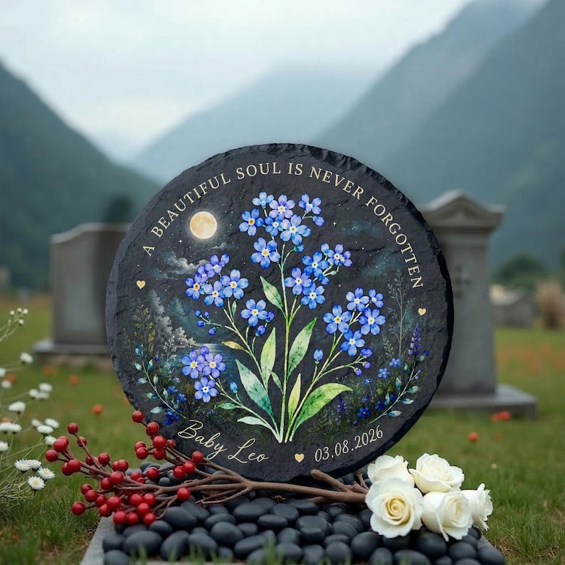 May include: A round, black memorial stone with a painted floral design of blue forget-me-nots. The text "A BEAUTIFUL SOUL IS NEVER FORGOTTEN" is arched around the top. The name "Baby Leo" and a date are at the bottom.