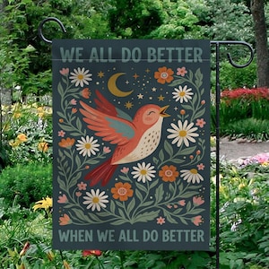 May include: A garden flag with the text "WE ALL DO BETTER WHEN WE ALL DO BETTER". The flag features a singing bird with orange and teal wings, surrounded by flowers, a crescent moon, and stars, all on a dark teal background.