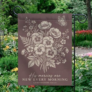 Op de afbeelding: Een bruine tuin vlag met een bloemenboeket en vlinder ontwerp. De vlag heeft de tekst "His mercies are NEW EVERY MORNING" en "LAMENTATIONS 3:22-23". De vlag hangt in een tuin.