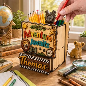 May include: A wooden desk organiser with the words "Adventure Awaits" and the name "Thomas". The organiser features a lion, giraffe, compass, and binoculars. Pencils and a compass are in the organiser. A small lion figurine is nearby.