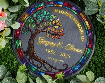 May include: A round, dark gray memorial stone with a stained-glass border. The design features a tree with colorful leaves and the inscription "In Loving Memory of Gregory E. Thomas 1952-2025."