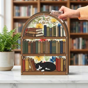 May include: A wooden bookshelf-shaped bank with the text "GRACE'S LIBRARY FUND". The bank features shelves filled with books and a sleeping black and white cat. Floral patterns decorate the background. A hand is inserting money into the slot.