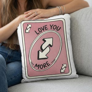 May include: A white pillow with a pink card design that says "LOVE YOU MORE". The design features a reverse arrow symbol in the center and in each corner. The pillow is on a gray couch.